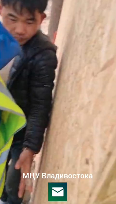 Video frame: close-up of a man in a black puffer jacket standing against a wall, beside a person in a yellow hi-vis vest at the left edge. Watermark: МЦУ Владивостока.