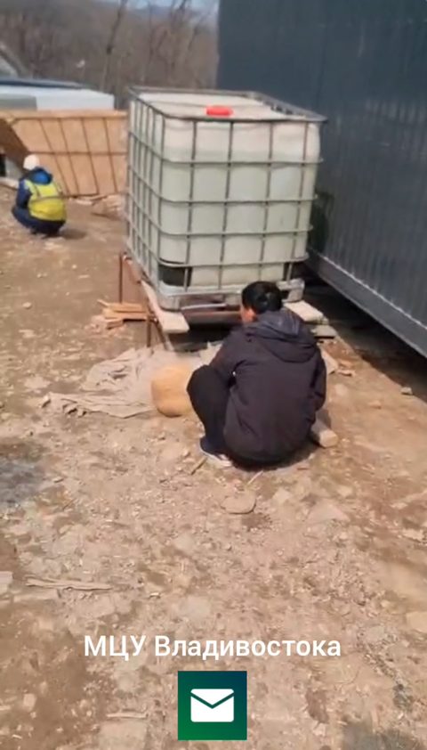 Video frame: a worker in a dark hooded jacket squats on bare dirt beside a white plastic IBC water tank on a pallet next to a grey metal container cabin; behind, a worker in a hi-vis vest and white hard hat crouches by a wooden fence, with bare trees on a hillside. Watermark: МЦУ Владивостока.