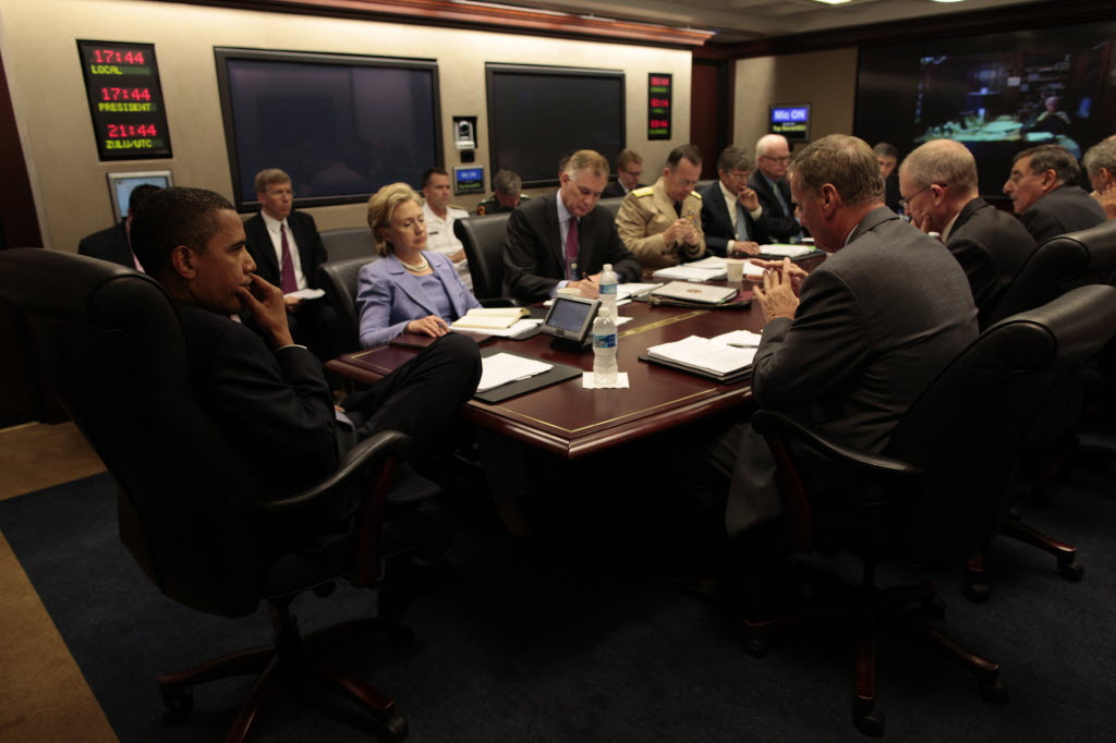 President Obama, seated in the foreground with his hand at his chin, faces Secretary of State Hillary Clinton and military and civilian officials around the Situation Room table, under clocks showing local, presidential and Zulu time.