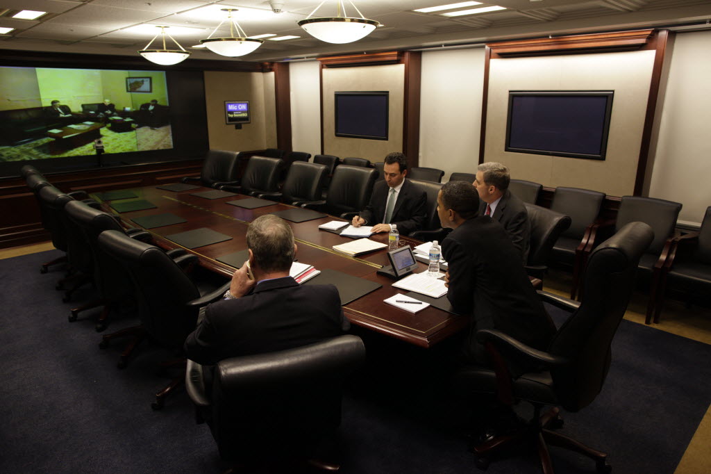 President Obama and three aides sit at the end of a long, mostly empty Situation Room table, facing a large wall screen that shows a room of people on the other end of the video link.