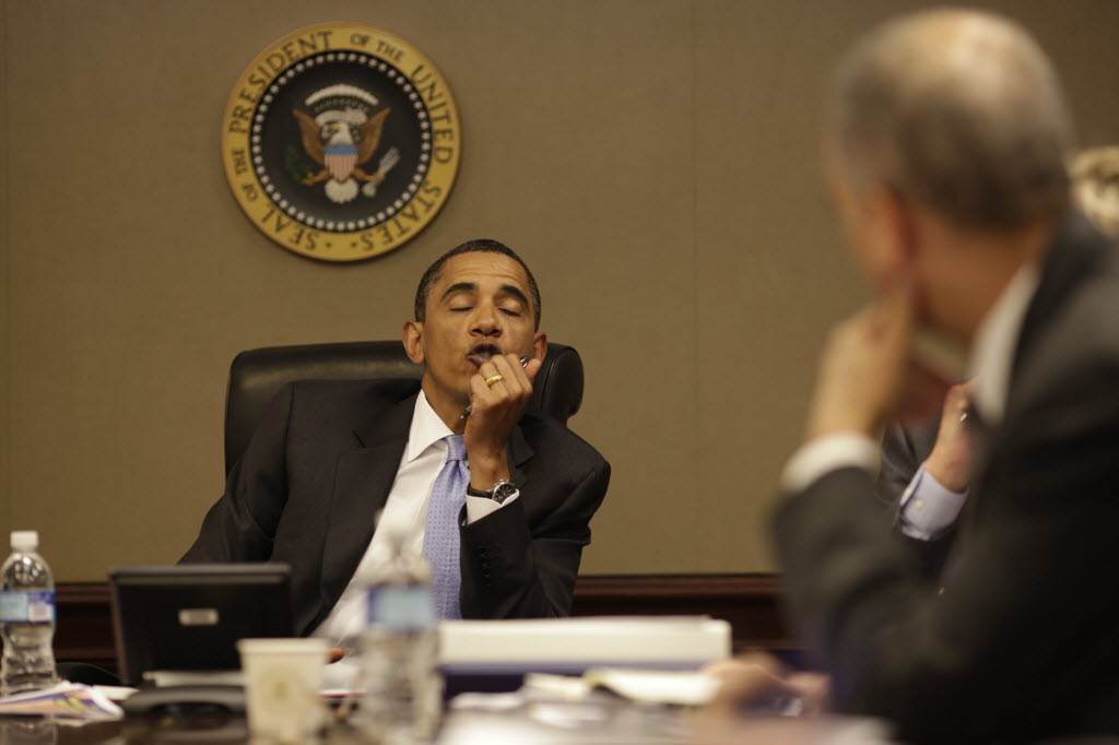 President Obama sits beneath the presidential seal with his chin on his hand, listening, as an official in the foreground turns toward him.