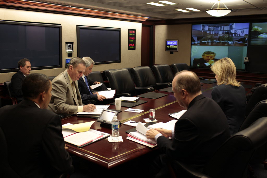 President Obama, in the foreground, and aides read papers at the Situation Room table while a wall screen at the far end shows participants on the video link, beneath a sign reading Mic On.