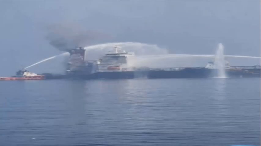 Wide shot of a long dark-hulled tanker with smoke rising from its aft superstructure while an orange tug at left and other vessels spray arcs of water onto it; a white superstructure stands beside the fire