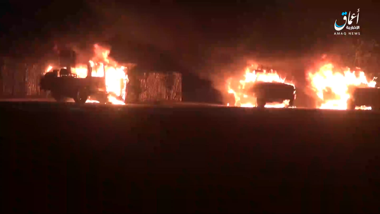 Burning vehicles at the base Nighttime scene of several military vehicles on fire beside corrugated-metal structures, with an Amaq News Agency watermark in the top-right corner