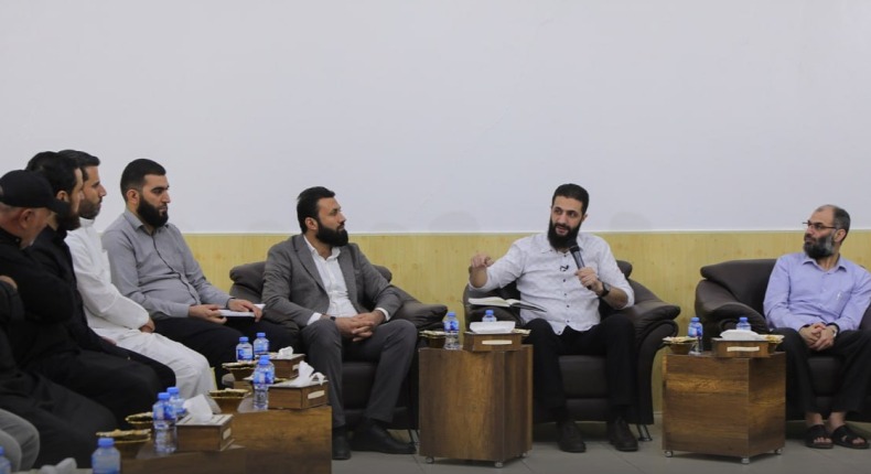 Al-Julani seated on a sofa between wooden side tables, talking with visitors in a white-walled hall, September 2023.