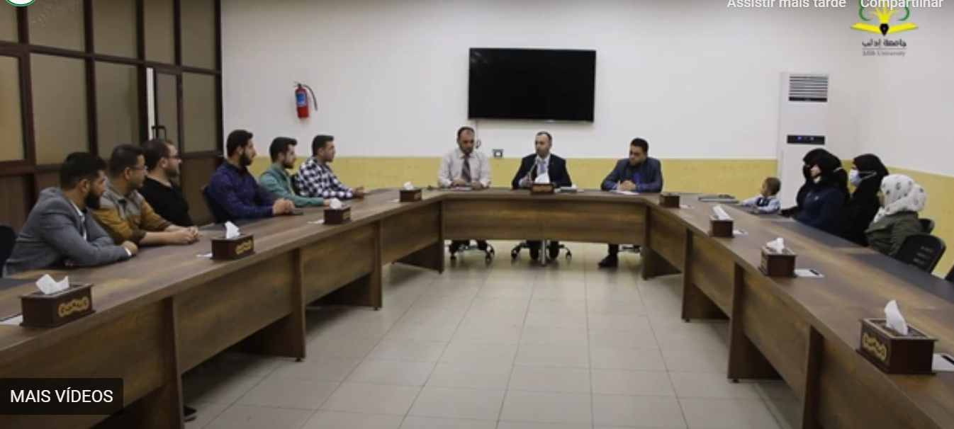 Idlib University students seated at long wooden tables in a hall at the Bab al-Hawa crossing, with a wall-mounted television and air conditioner.