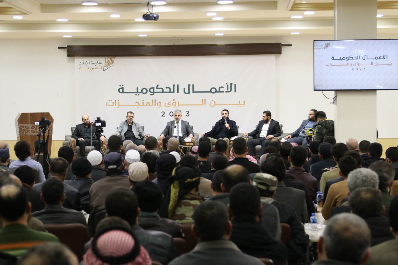 Al-Julani on stage at a Salvation Government symposium under a banner reading Government Work Between Visions and Achievements 2023.