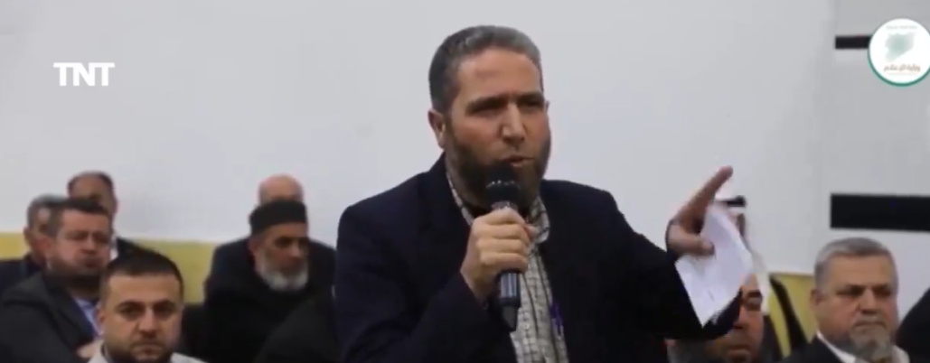 Video frame from the same event: a man speaking into a microphone in front of light wood wall paneling.