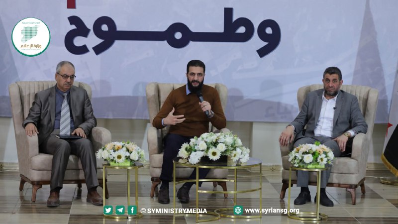 Al-Julani seated between two men in armchairs on a stage decorated with flowers, March 30, 2024.